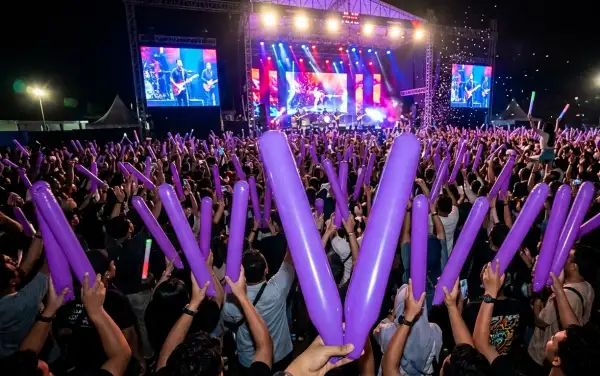 Penonton mengangkat balon tepuk berwarna ungu untuk memeriahkan suasana konser musik pada malam hari.