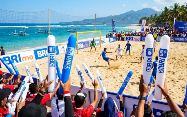 Penonton mengangkat balon tepuk berlogo sponsor saat pertandingan sepak bola pantai berlangsung.