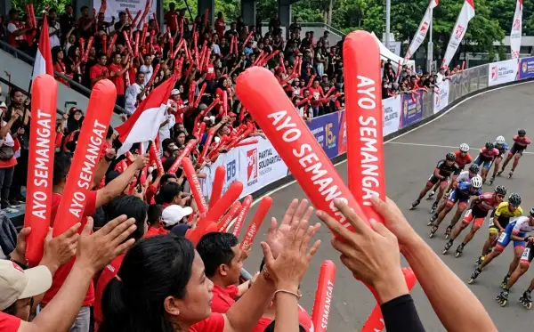 Suporter mengangkat balon tepuk merah sambil menyaksikan perlombaan sepatu roda di lintasan.